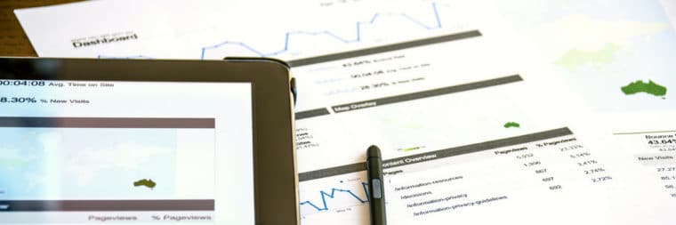 A close-up view of a laptop displaying data and maps. A paper with data, numbers, and graphs is also visible, along with a pen.