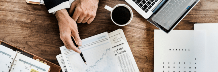 From a top-down perspective, the hands of a man are visible holding a pen, while next to him is a laptop, a cup of coffee, a newspaper, a calendar, and an agenda.