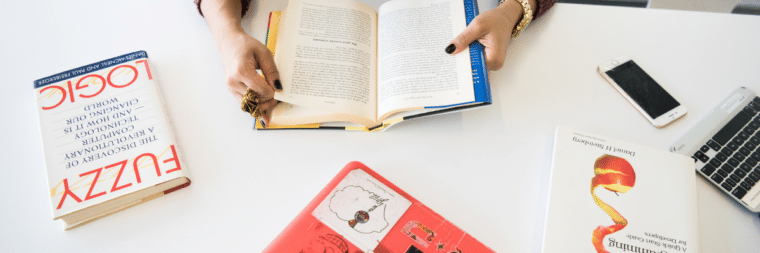 On a table, there are several books. A woman's hands are visible as she hands one of the books. There is also a phone and a laptop on the table.