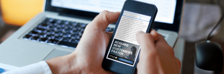 Someone's hand holding a phone displaying the MRP website, with the visible text 'Acquire new customers faster.' In the background, a laptop keyboard is visible.